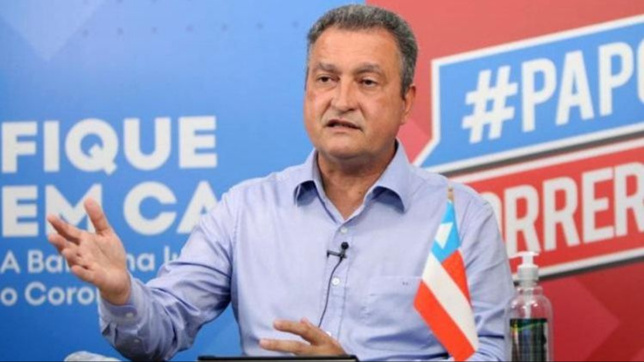 Foto do governador da Bahia, Rui Costa, gesticulando com os braços. ele está usando uma camisa de cor clara e ao fundo há um painel com palavras pedindo que as pessoas fiquem em casa.