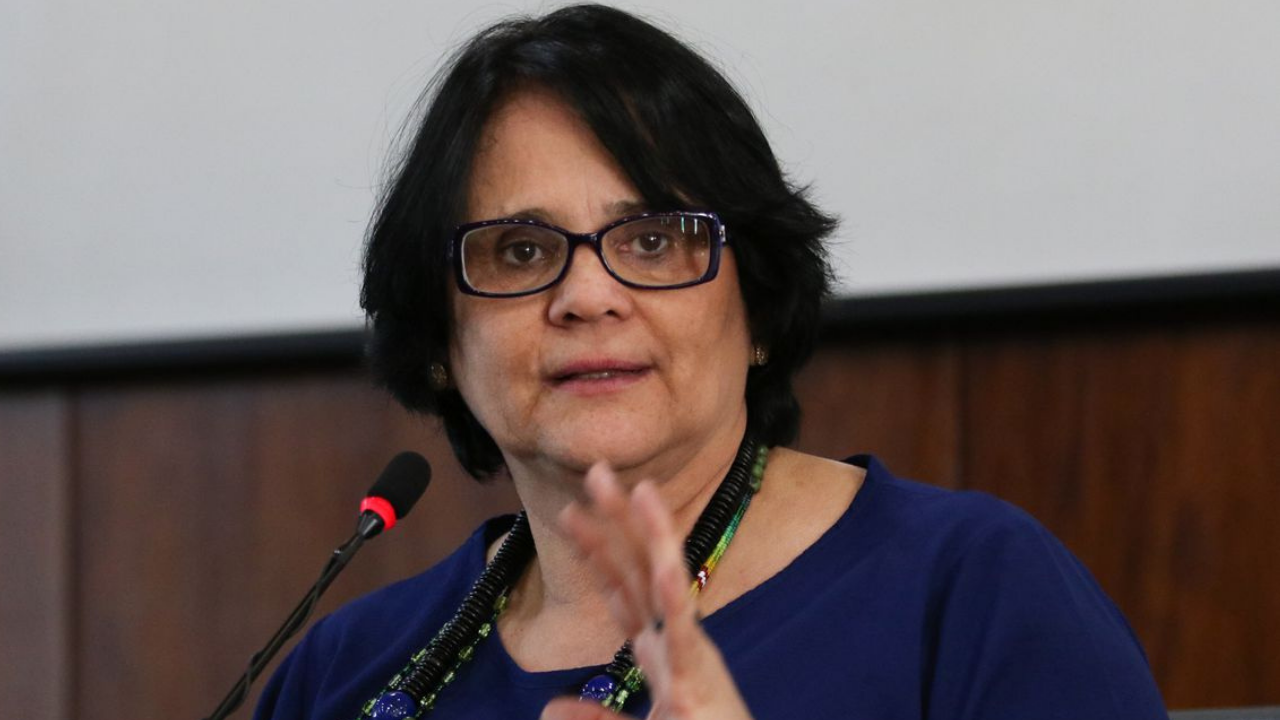 Foto da Ministra Damares Alves falando so microfone. Ela usa uma blusa verde e óculos de grau.