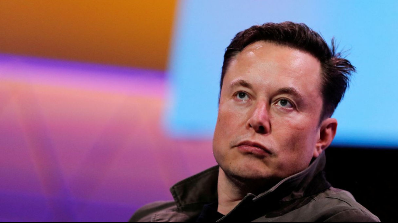 Elon Musk com olhar desatento e cara séria. Ele é branco e usa cabelo preto, fundo colorido desfocado.