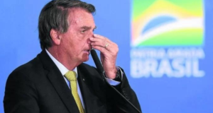 Bolsonaro fala ao microfone com mão direita segurando o nariz.