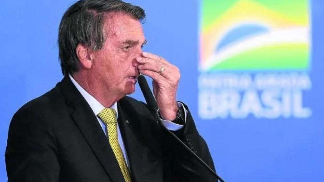 Bolsonaro fala ao microfone com mão direita segurando o nariz.