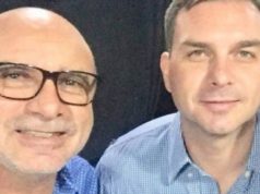 Foto de Fabricio Queiroz, à esquerda, e Flávio Bolsonaro à direita. Eles posam para selfie com olhar sério portando camisas azuis. Cor preta ao fundo.