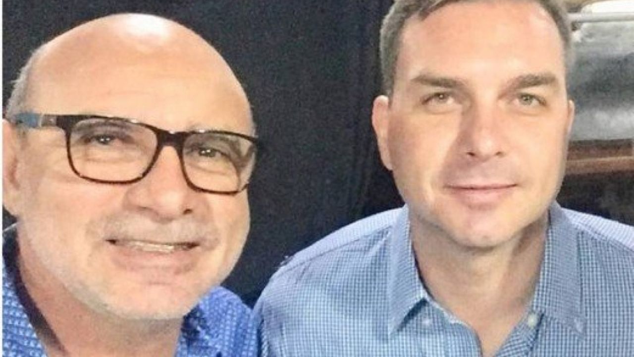 Foto de Fabricio Queiroz, à esquerda, e Flávio Bolsonaro à direita. Eles posam para selfie com olhar sério portando camisas azuis. Cor preta ao fundo.