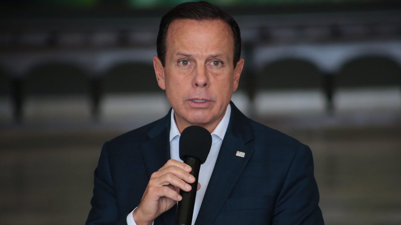 Foto de Joao Doria flando ao microfone. Ele está sem barba, sem máscara, tem cabelo preto e usa terno de cor azul marinho.