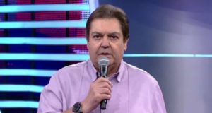 Faustão com o microfone na mão direita posicionado na boca, camisa rosa e olhar sério.