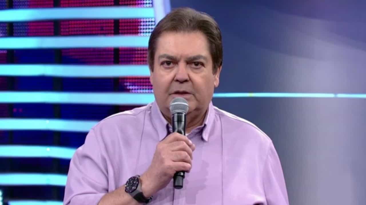 Faustão com o microfone na mão direita posicionado na boca, camisa rosa e olhar sério.