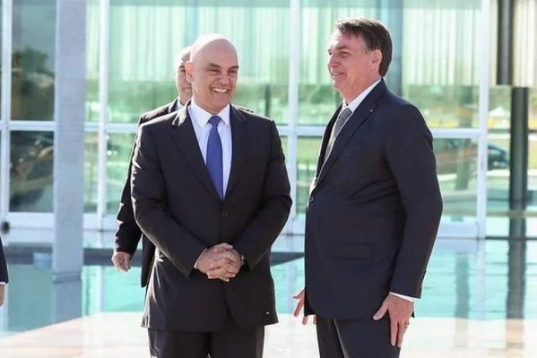 O ministro do STF Alexandre de Moraes ao lado do presidente Jair Bolsonaro (PL), ambos de pé e sorrindo.