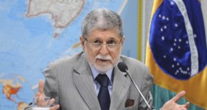 Celso Amorim defende chapa Lula Alckmin. Ele está em uma mesa, conversando com alguém. Usa óculos e tem cabelo grisalho, veste terno cinza.