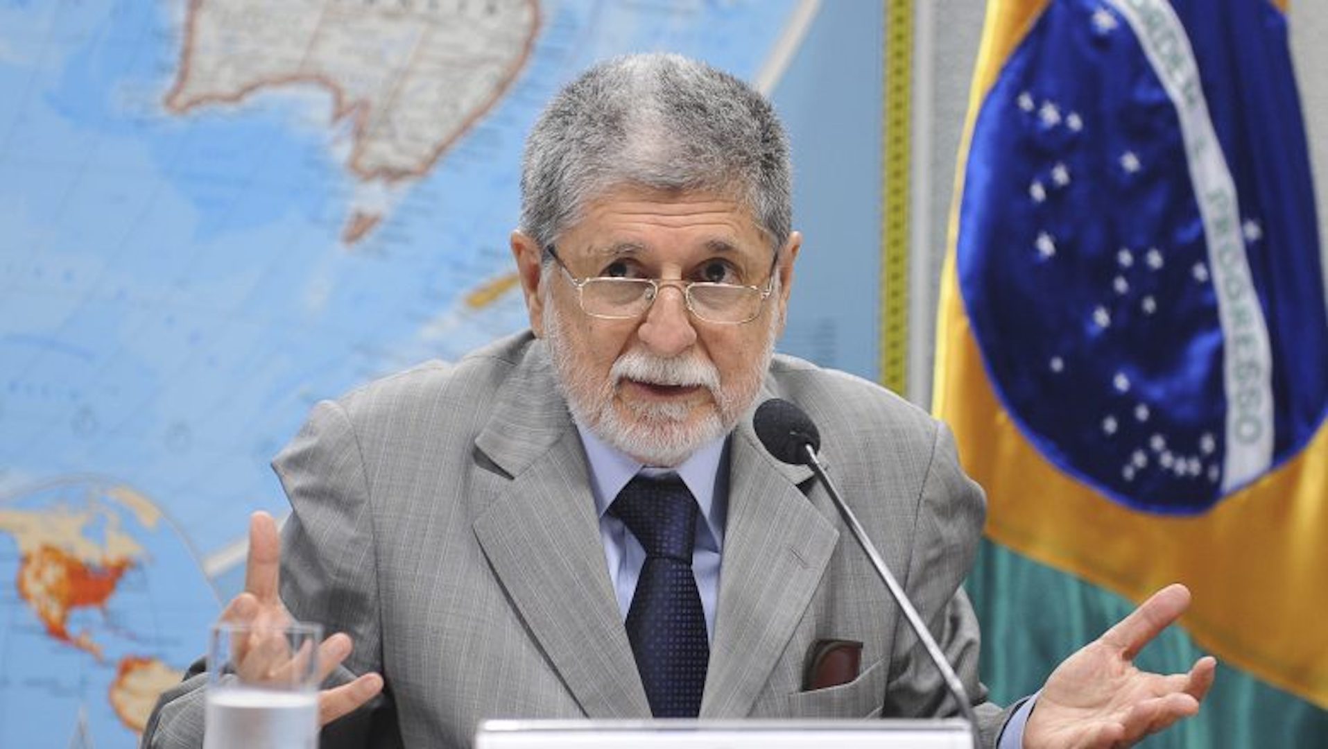 Celso Amorim defende chapa Lula Alckmin. Ele está em uma mesa, conversando com alguém. Usa óculos e tem cabelo grisalho, veste terno cinza.