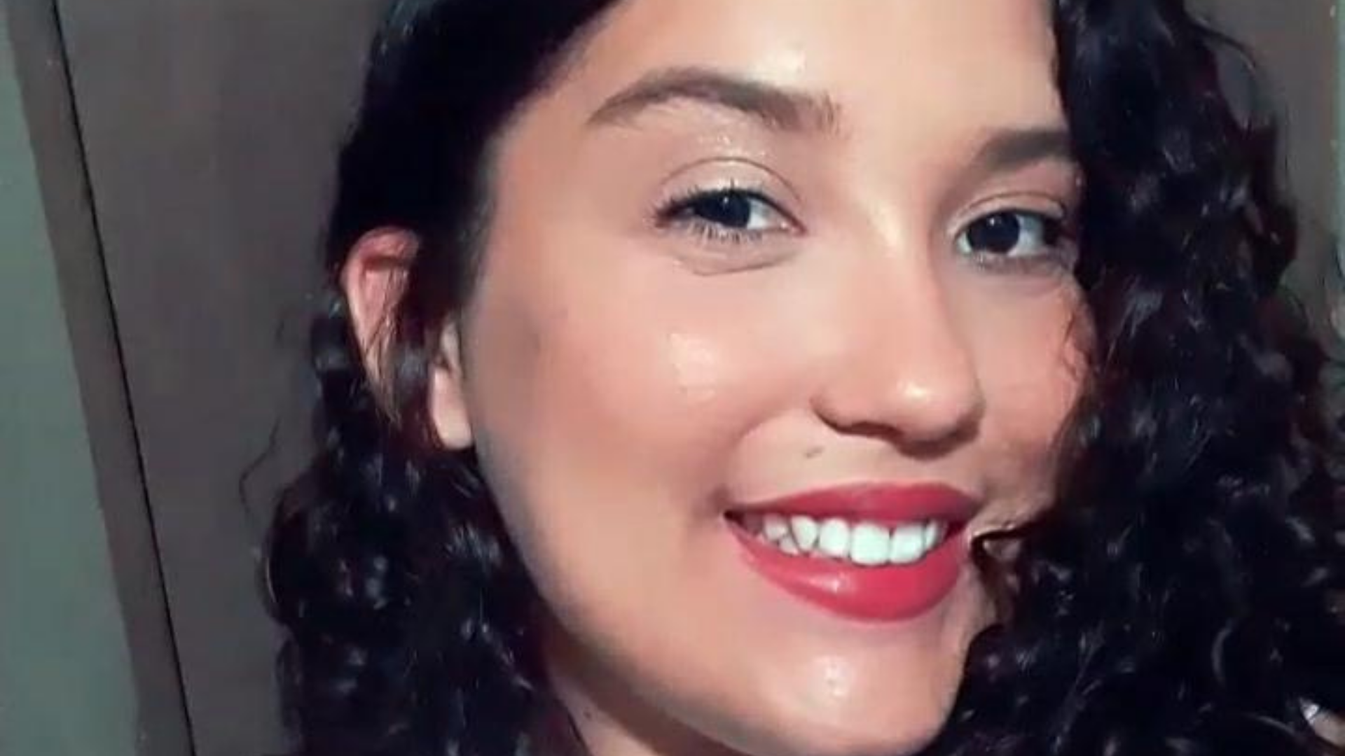 Milena Vieira usa cabelo preto cahceado, batom vermelho e está sorrindo na foto.