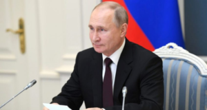 Foto de Putin em uma cadeira, assinando um documento. Ele usa terno preto, tem pele branca e é calvo. O fundo está desfocado.