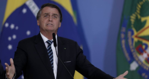 Homem ameaçou colocar veneno em comida de Bolsonaro. Presidente está com expressão indignada, falando ao microfone com símbolo do brasil ao fundo