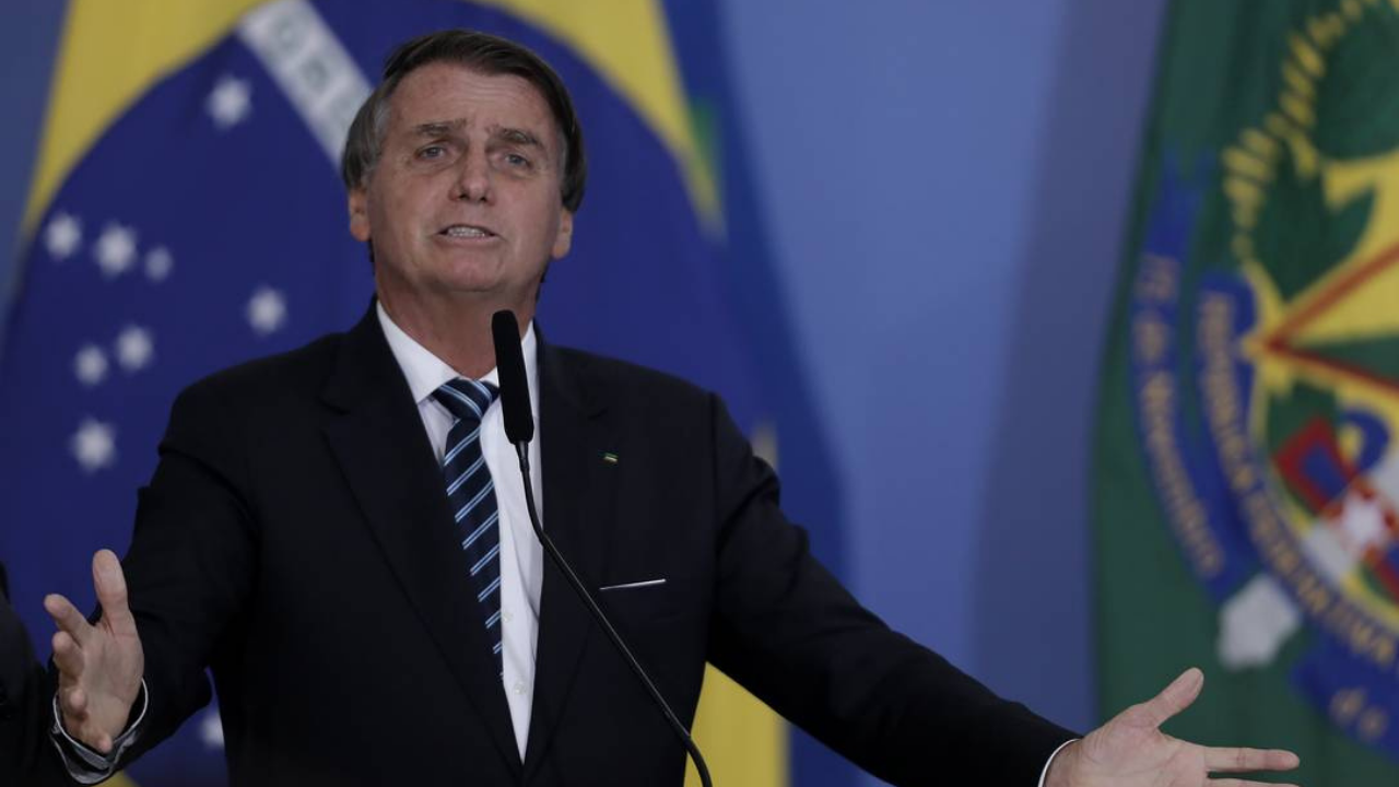Homem ameaçou colocar veneno em comida de Bolsonaro. Presidente está com expressão indignada, falando ao microfone com símbolo do brasil ao fundo