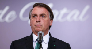 Bolsonaro usando terno azul e falando em microfone. Há um fundo rosa.