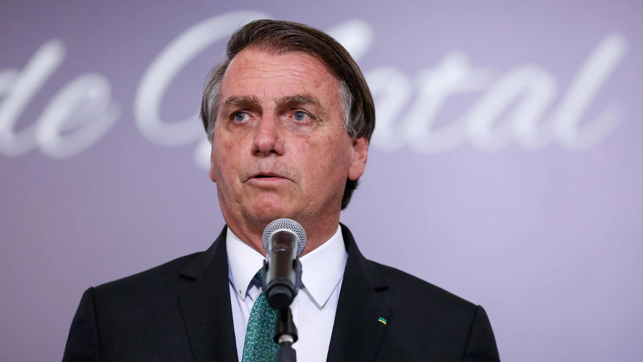 Bolsonaro usando terno azul e falando em microfone. Há um fundo rosa.