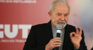 Foto de lula falando ao microfone. Ele usa terno preto. Ao fundo há um painel da CUT (Central Única dos Trabalhadores)