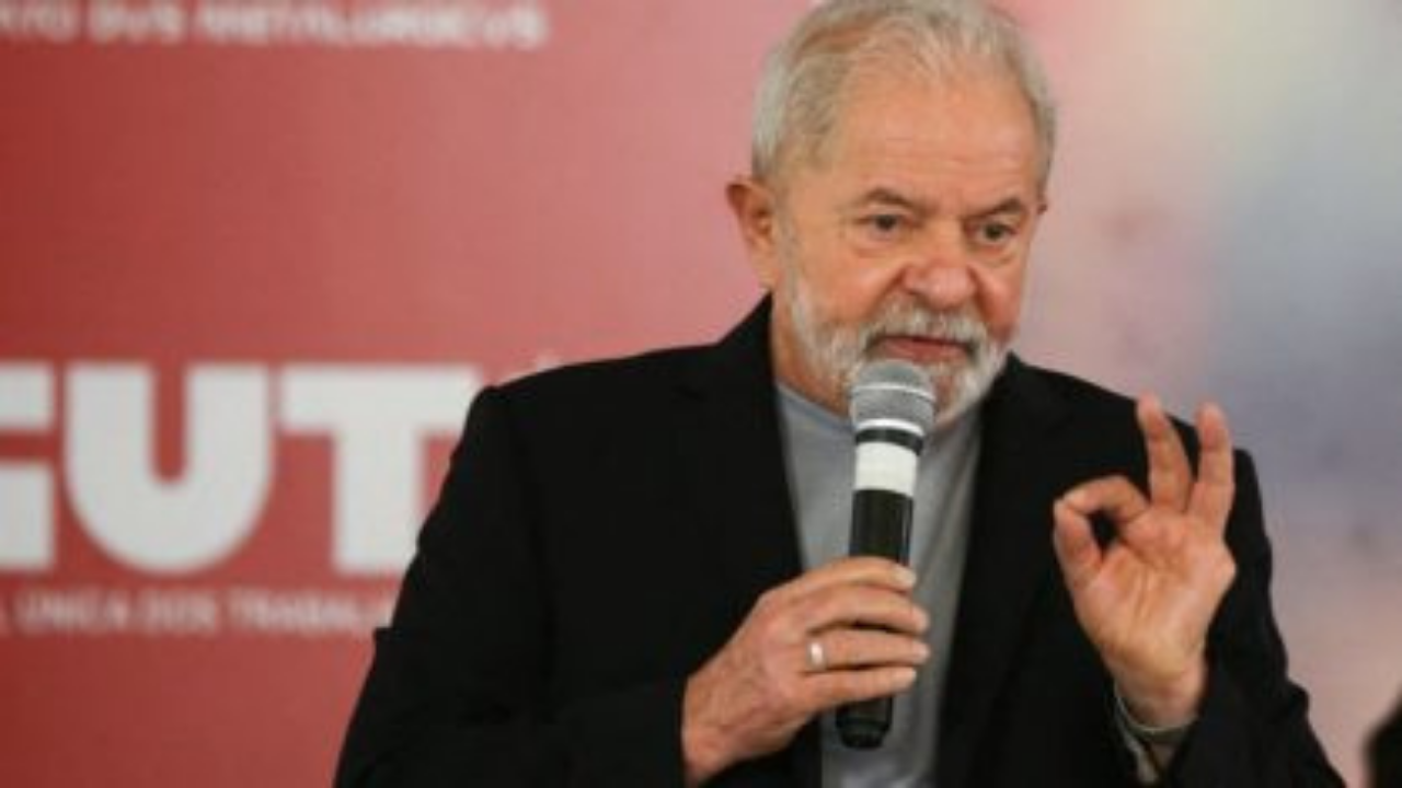 Foto de lula falando ao microfone. Ele usa terno preto. Ao fundo há um painel da CUT (Central Única dos Trabalhadores)