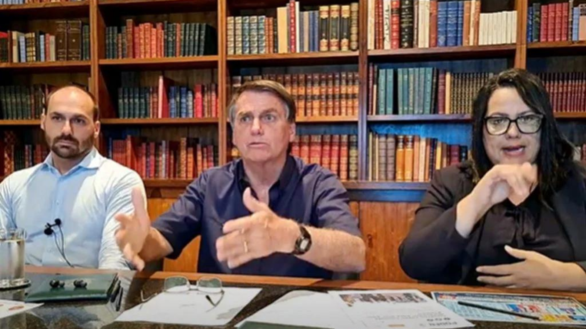 . Bolsonaro afirmou que o Brasil teria passado por lockdown nacional se o presidente fosse Haddad. Ele aparece na foto com Eduardo Boslonaro, à equerda, e intérprete de libras, à direita. Ao fundo há uma estante com livres.