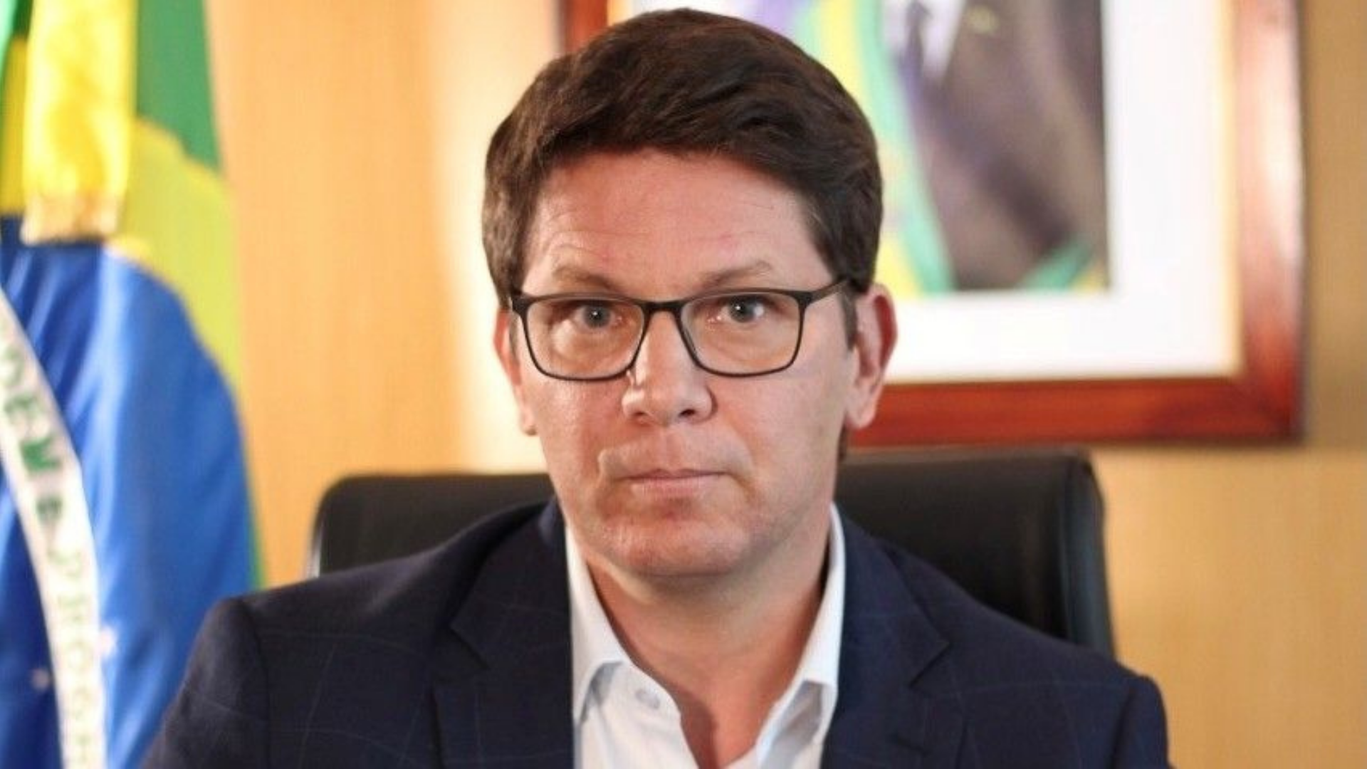 Mário Frias sofre dura derrota. Na foto, ele tem olhar sério, usa óculos, cabelo preto e ao fundo há uma bandeira do Brasile foto do presidente Bolsonaro.