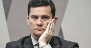 Moro em pânico para conseguir recursos financeiros. Ele usa terno preto, tem a mão esquerda na bochecha e olhar sério.