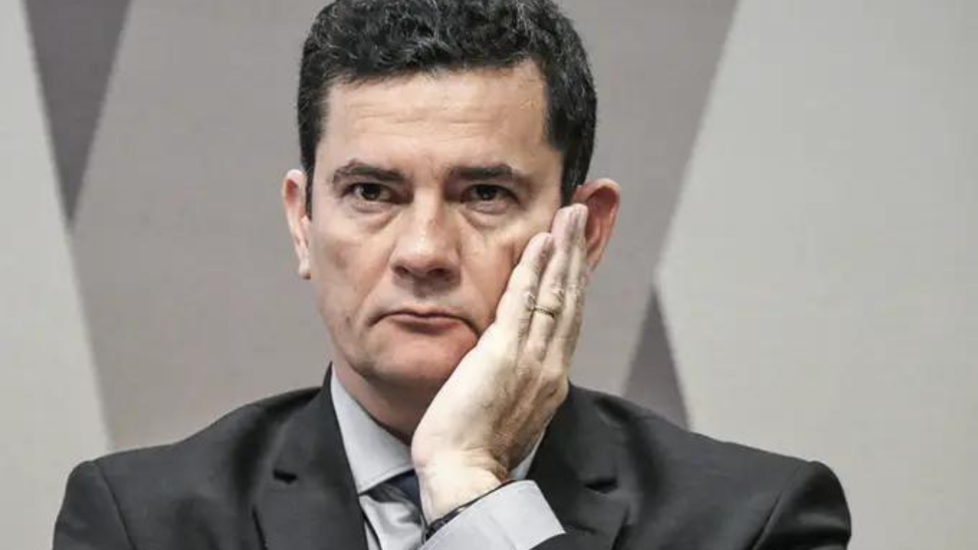 Moro em pânico para conseguir recursos financeiros. Ele usa terno preto, tem a mão esquerda na bochecha e olhar sério.