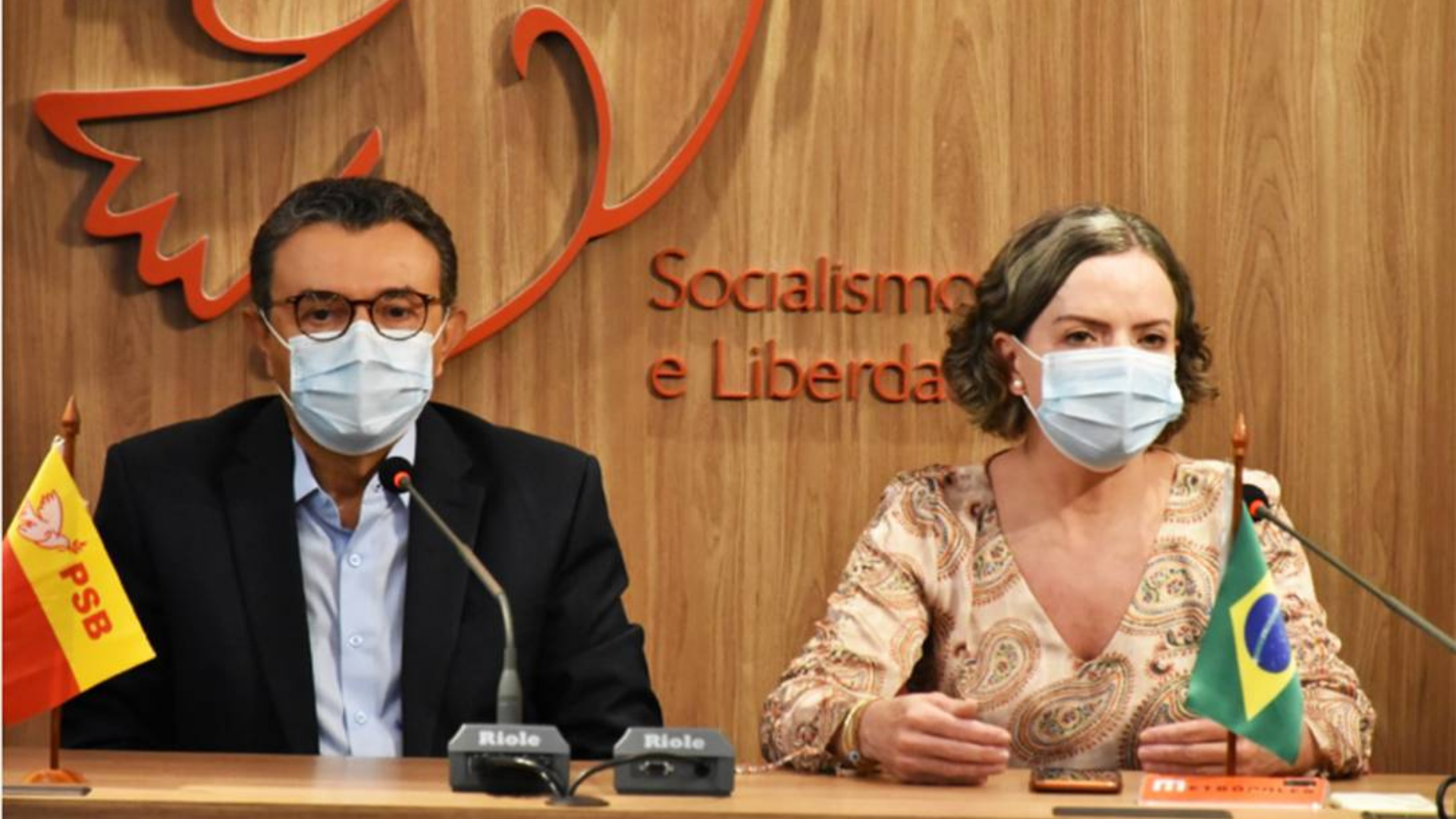 Gleisi detona governador do PSB. Ela está em uma mesa com o presidente do PSB (à esquerda). Ambos usam máscara e ao fundo há a logo do PSB.