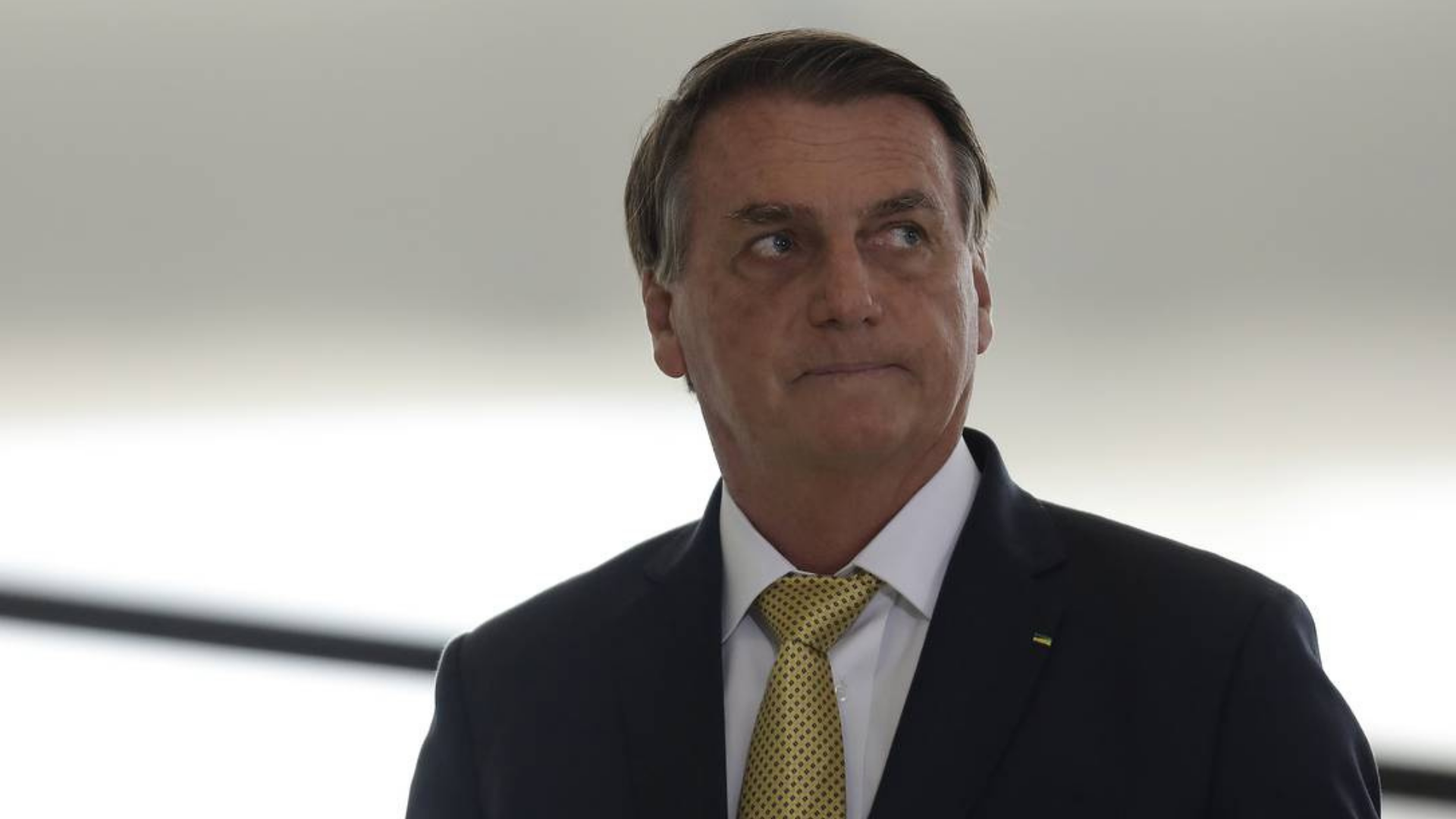 Segurança da equipe de Bolsonaro é flagrado agredindo jovens. ele usa terno cinza e gravata amarela, olhar confuso com a cabeça virada para a esquerda.