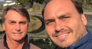 Bolsonaro em selfie com o filho Carlos Bolsonaro. Eles tem pele branca, e cabelos castanhos. Usam casacos para o fio, mas o dia está ensolarado.