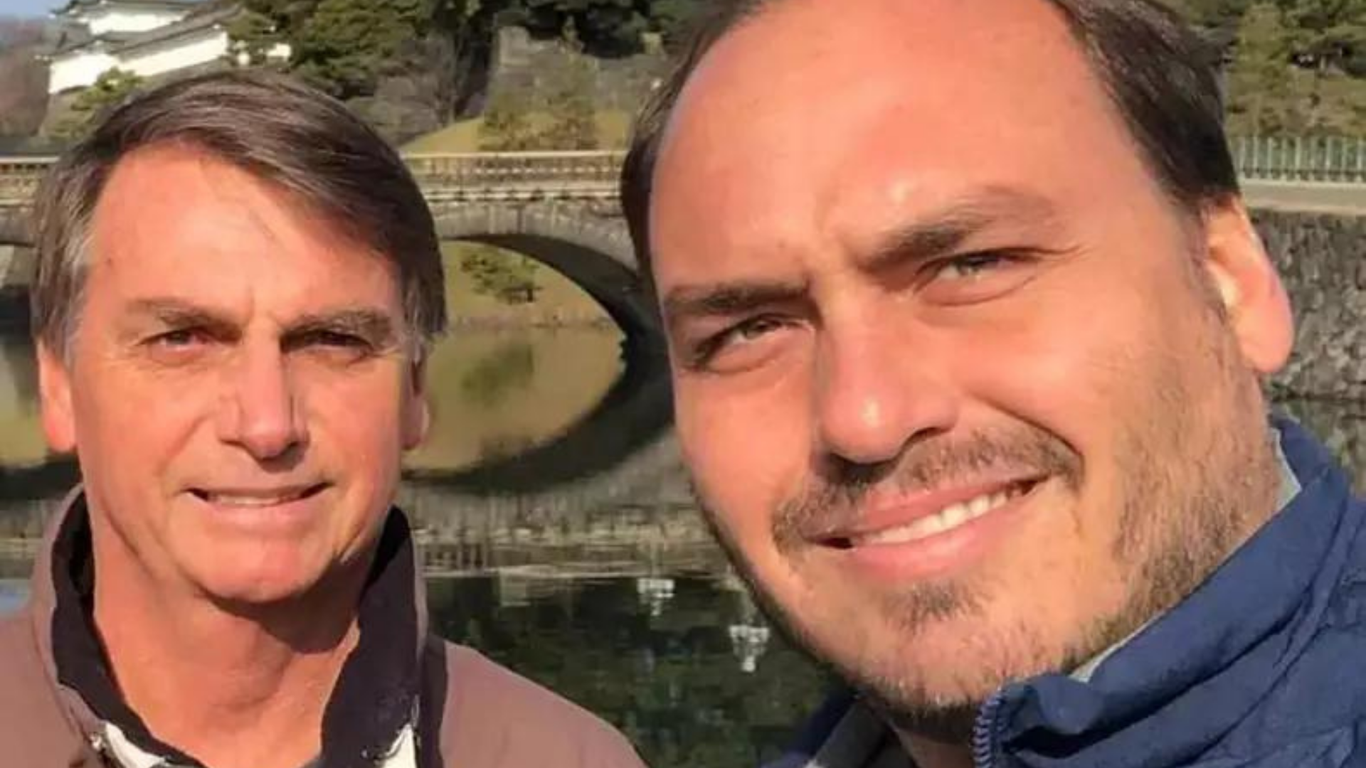 Bolsonaro humilha ministros para defender Carluxo na Rússia. Ele está em selfie com o filho Carlos Bolsonaro. Eles tem pele branca, e cabelos castanhos. Usam casacos para o fio, mas o dia está ensolarado.