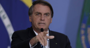 O presidente Jair Bolsonaro elogia a economia. Ele aparece em foto com o indicador apontado, verbalizando algo. Com um fundo azul. ele usa terno preto.