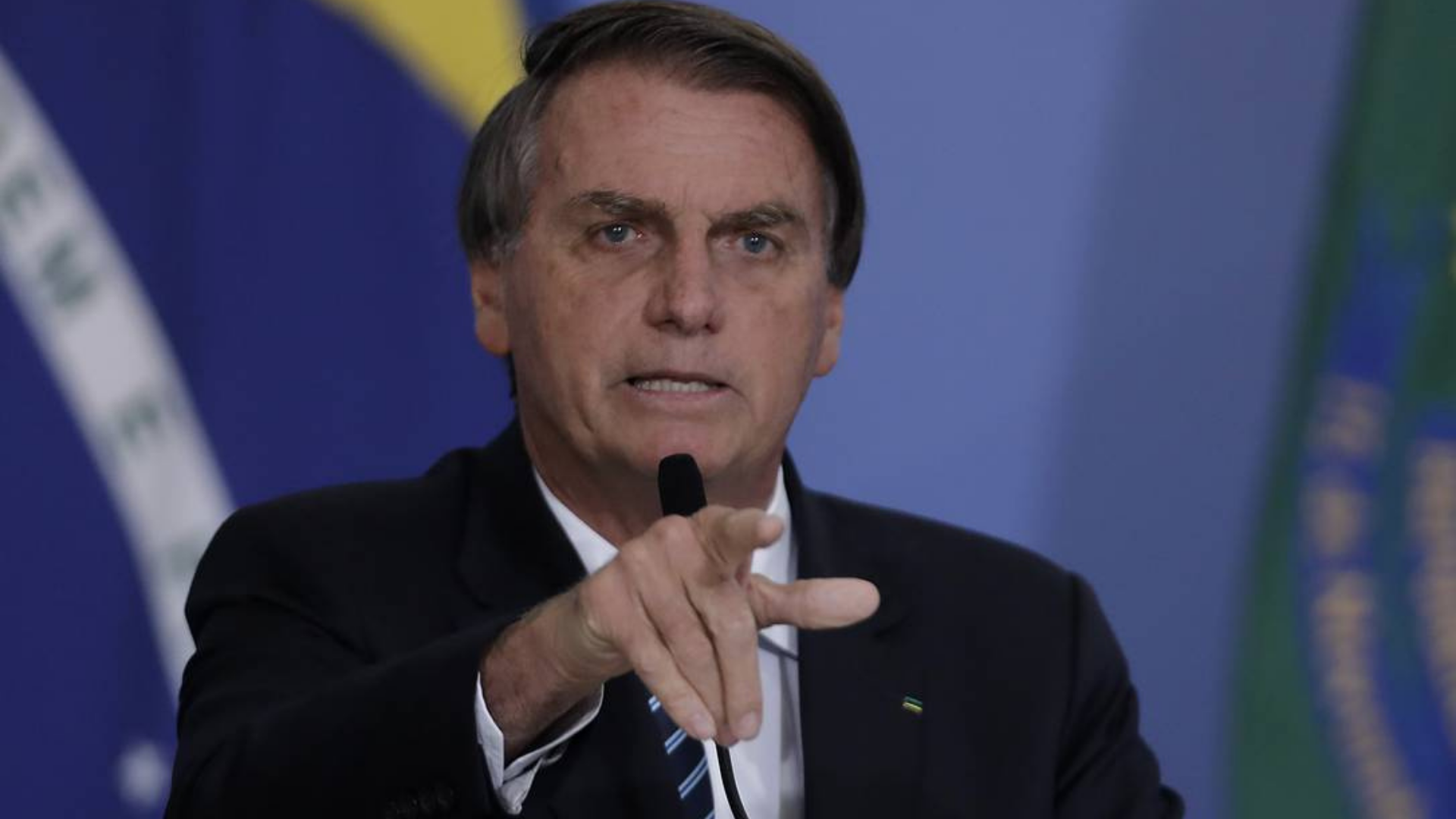 O presidente Jair Bolsonaro elogia a economia. Ele aparece em foto com o indicador apontado, verbalizando algo. Com um fundo azul. ele usa terno preto.