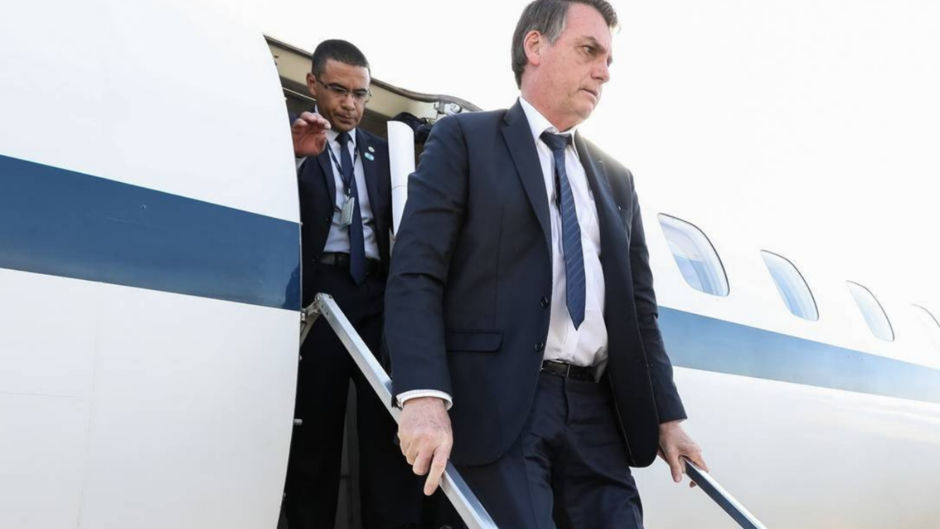 Após fracasso em viagem à Europa, Bolsonaro promete visita à América Central. Ele usa terno preto e gravata azul, está descendo de um avião.