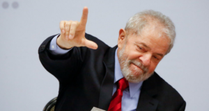 Lula usa terno preto e aponta a mão direita pra cima em formato de L.