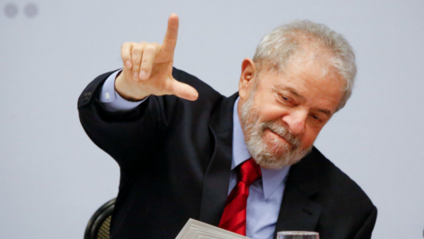 Lula usa terno preto e aponta a mão direita pra cima em formato de L.