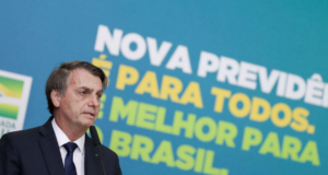 Foto de Bosonaro falando ao microfone. Ele us aum terno azul, ao fundo há um painel com frases sobre a reforma da previdência.