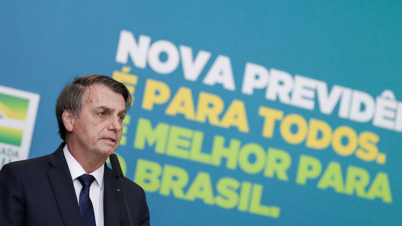 Foto de Bosonaro falando ao microfone. Ele us aum terno azul, ao fundo há um painel com frases sobre a reforma da previdência.