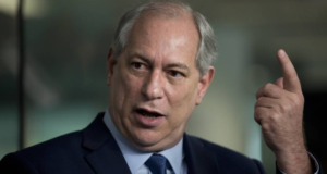 Ciro Gomes é desmascarado após postar foto antiga. Ele usa terno preto e faz sinal de susto com os olhos, levantando o indicado direito para cima.