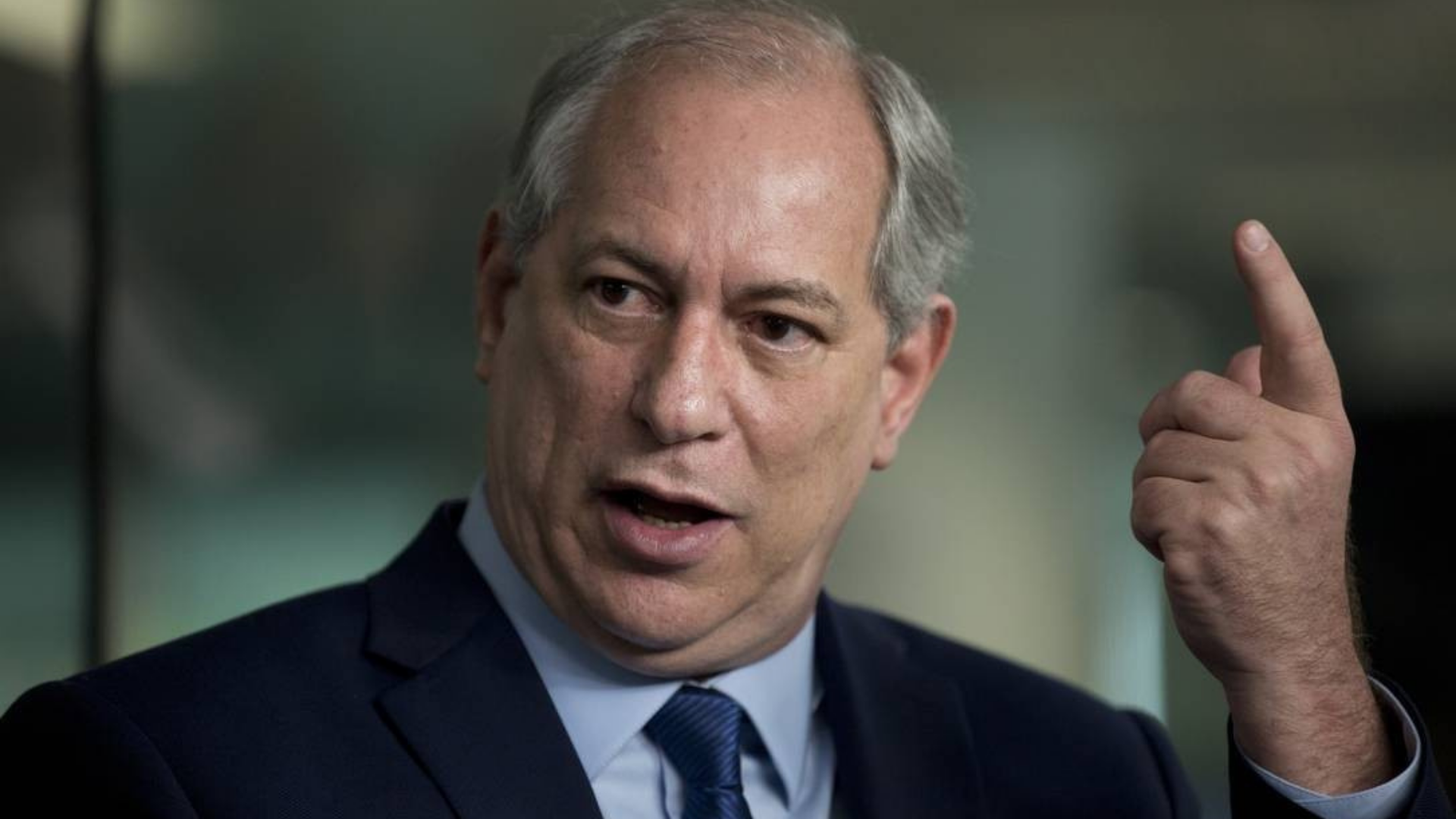 Ciro Gomes é desmascarado após postar foto antiga. Ele usa terno preto e faz sinal de susto com os olhos, levantando o indicado direito para cima.