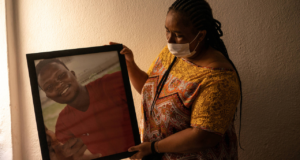 Foto da mãe de Moise, ela é negra, tem tranças no cabelo, usa mascara, uma blusa marrom e segura uma fotografia de Moise com as duas maos.