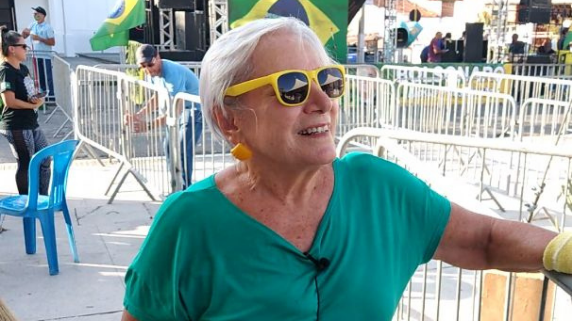 Mirna usa blusa verde, um óculos amarelo e um sorriso no rosto. Ela tem pelo branca e cabelo loiro.