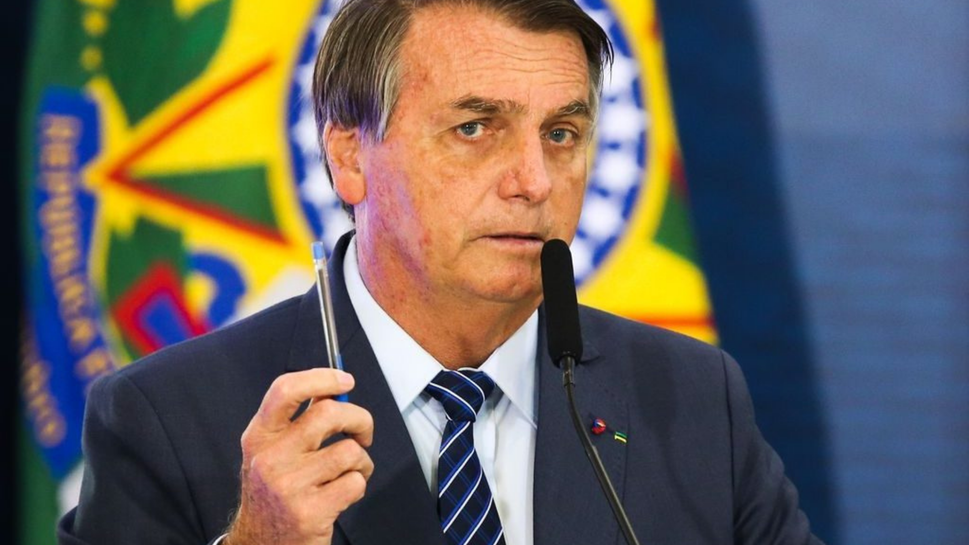 Bolsonaro aparece usando um terno preto, com gravata azul e listras brancas. Ele segura uma caneta azul com a mão direita.