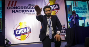 Brasileiros se filiam a partido de extrema-direita em Portugal. O candidato levanta a mão direita, usa terno preto e painel com logo do partido ao fundo.