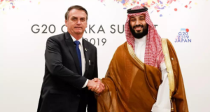 Bolsonaro convida príncipe saudita que mandou esquartejar jornalista. Ele está fazendo um aperto de mãos com o príncipe, que usa trajes tradicionais do país. Bolsonaro usa terno preto e gravata azul.