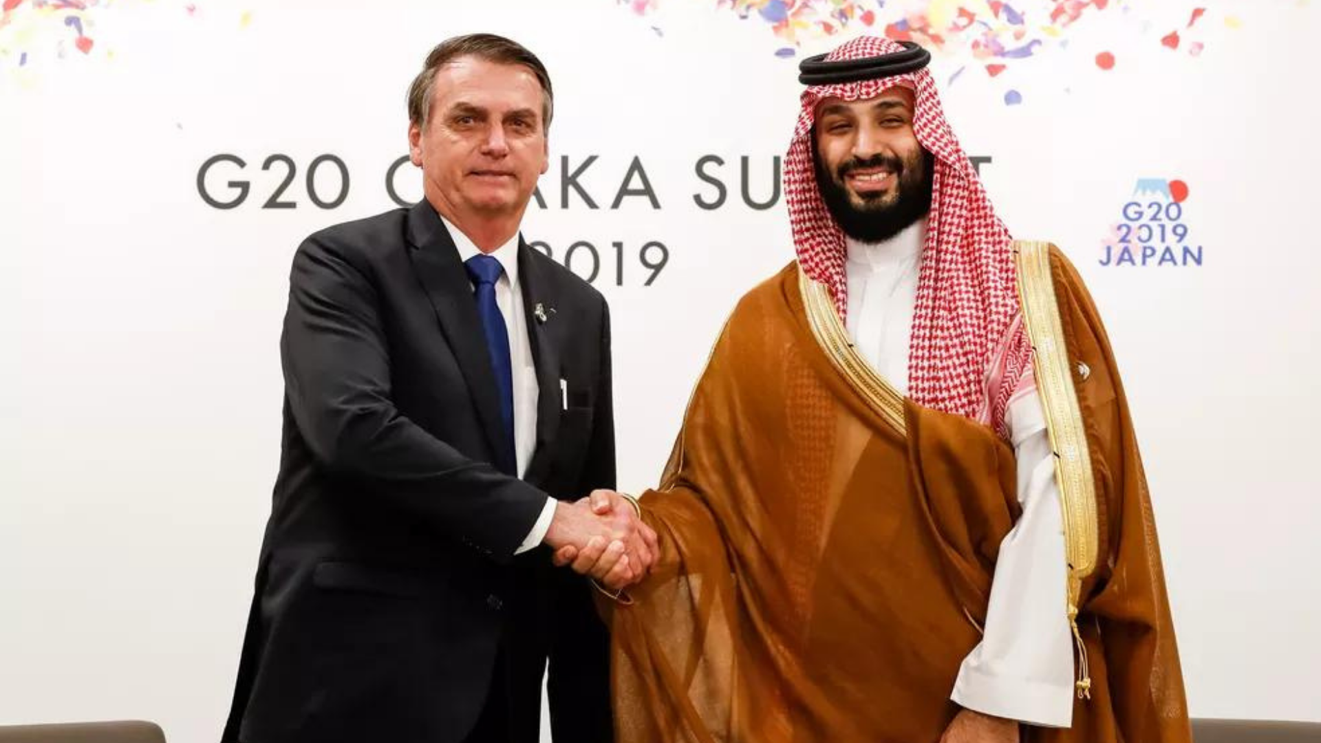 Bolsonaro convida príncipe saudita que mandou esquartejar jornalista. Ele está fazendo um aperto de mãos com o príncipe, que usa trajes tradicionais do país. Bolsonaro usa terno preto e gravata azul.