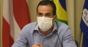 Prefeito de Salvador com camisa azul e branco, usando máscara branca e com olhar sério. Ele não usa barba e tem cabelos pretos.