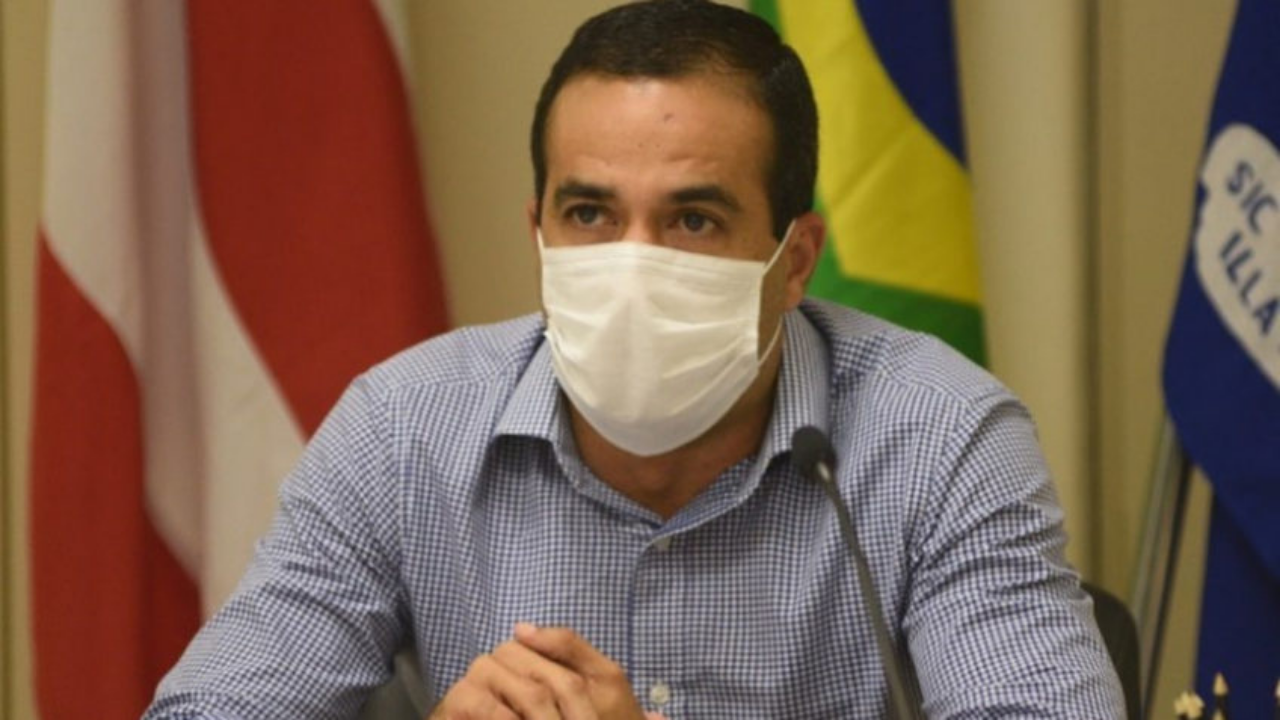 Prefeito de Salvador com camisa azul e branco, usando máscara branca e com olhar sério. Ele não usa barba e tem cabelos pretos.