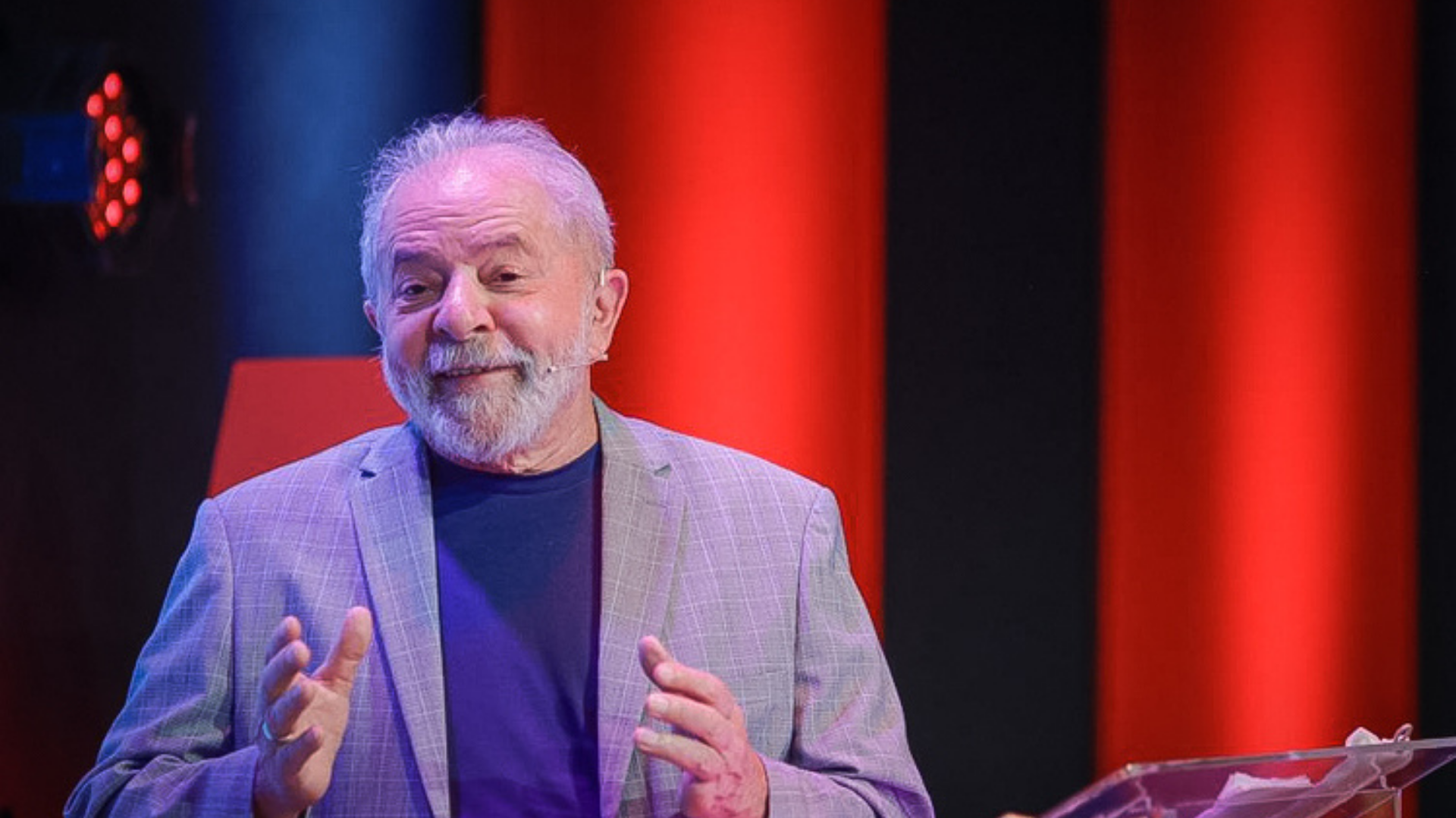 Lula relembra momentos da história do PT em live. Ele usa terno, sorri para a câmera e há um fundo vermelho no local.