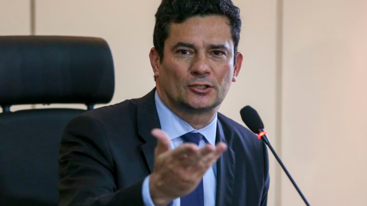 Foto de Sergio Moro usando terno preto e gravada azul. Ele não tem barba e fala ao microfonene