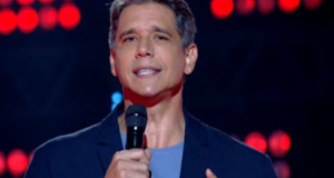 Márcio Garcia condenado a pagar multa por atrasar dúvida. Ele usa roupas em tom azul e fala ao microfone durante programa de TV.