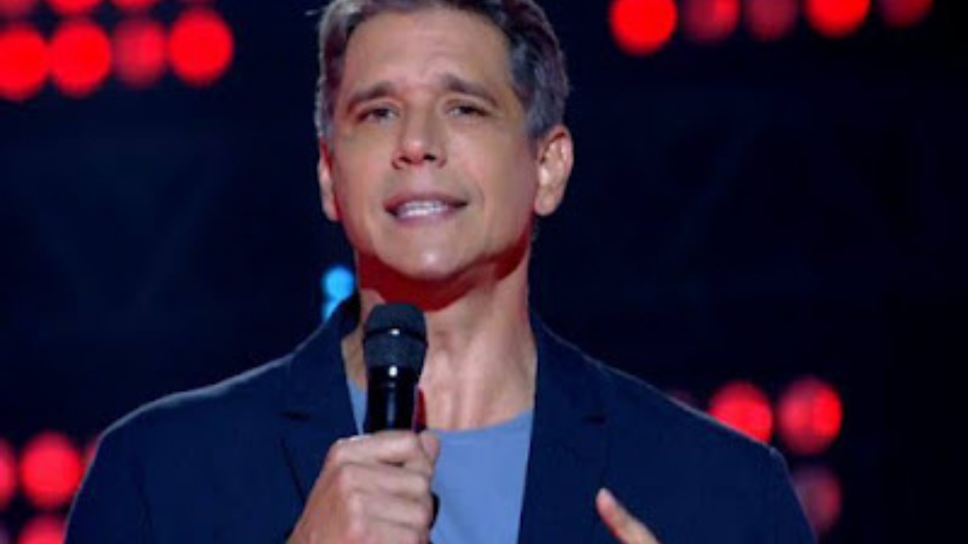 Márcio Garcia condenado a pagar multa por atrasar dúvida. Ele usa roupas em tom azul e fala ao microfone durante programa de TV.